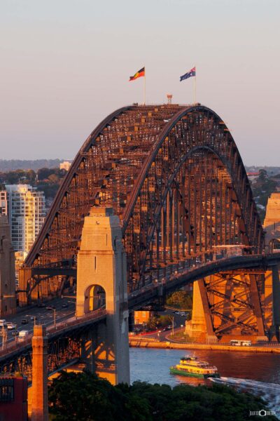 Vertical photograph of the Sydney Harbour Bridge glowing in warm evening sunset light over Sydney Harbour.