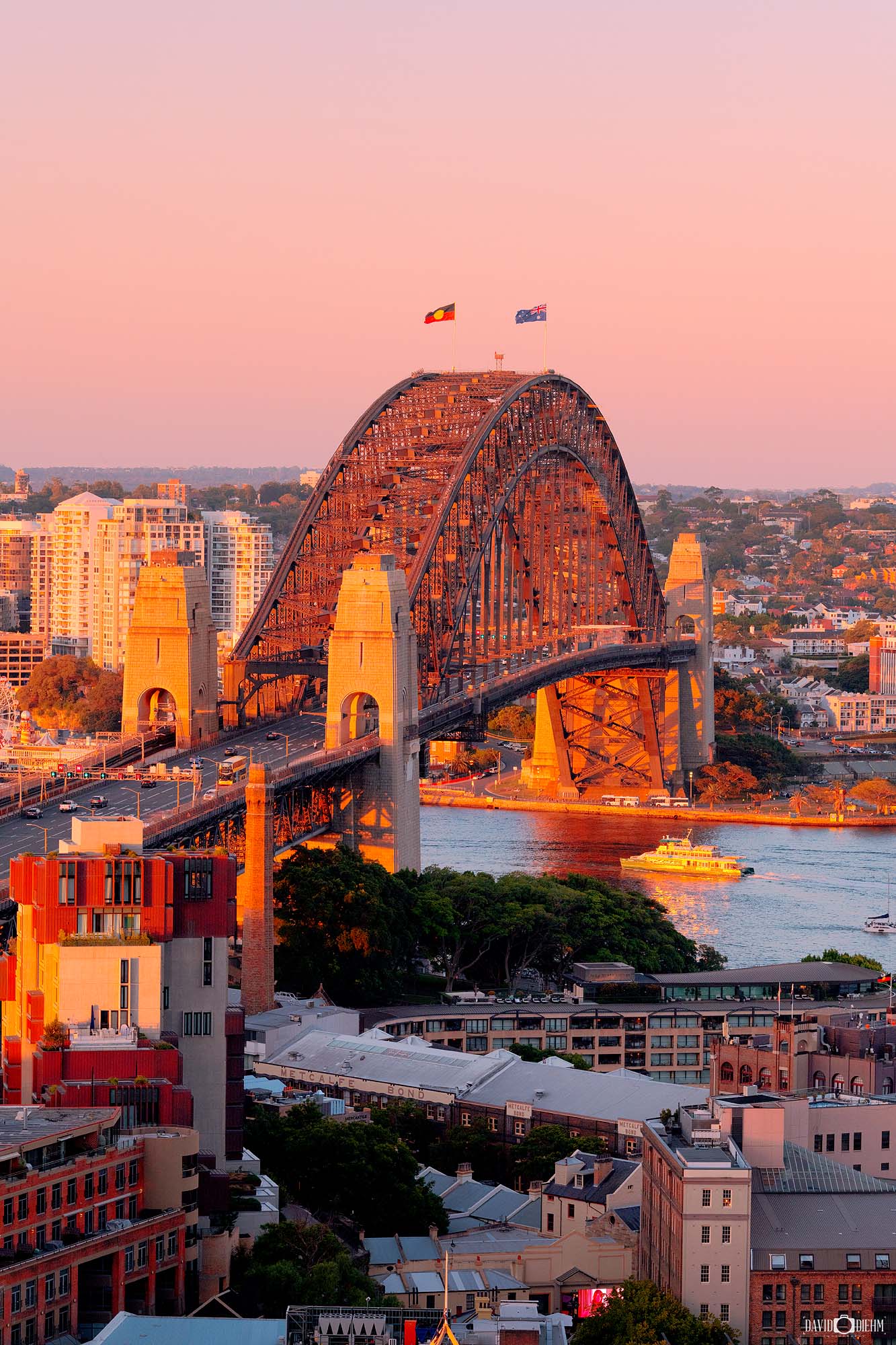 Portrait-format photograph of the Sydney Harbour Bridge in warm sunset tones with glowing skies over Sydney Harbour.
