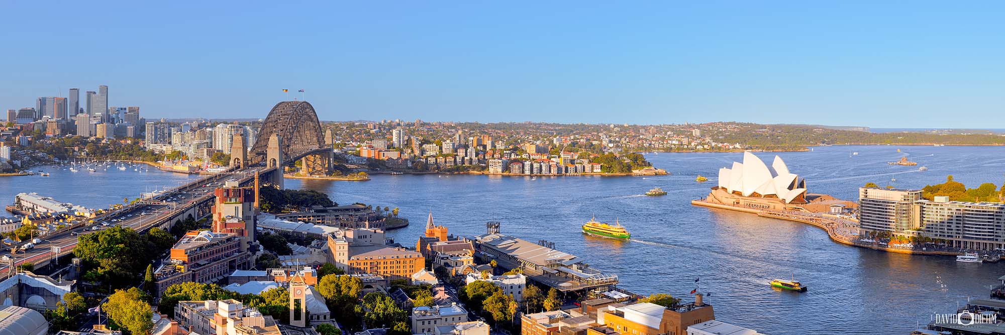 Panoramic photograph of Sydney Harbour during the day featuring blue water, city skyline, and iconic waterfront landmarks under clear skies.