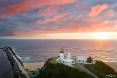 Aerial view of Nobbys Lighthouse in Newcastle at sunrise with warm morning light over the coastline and breakwall