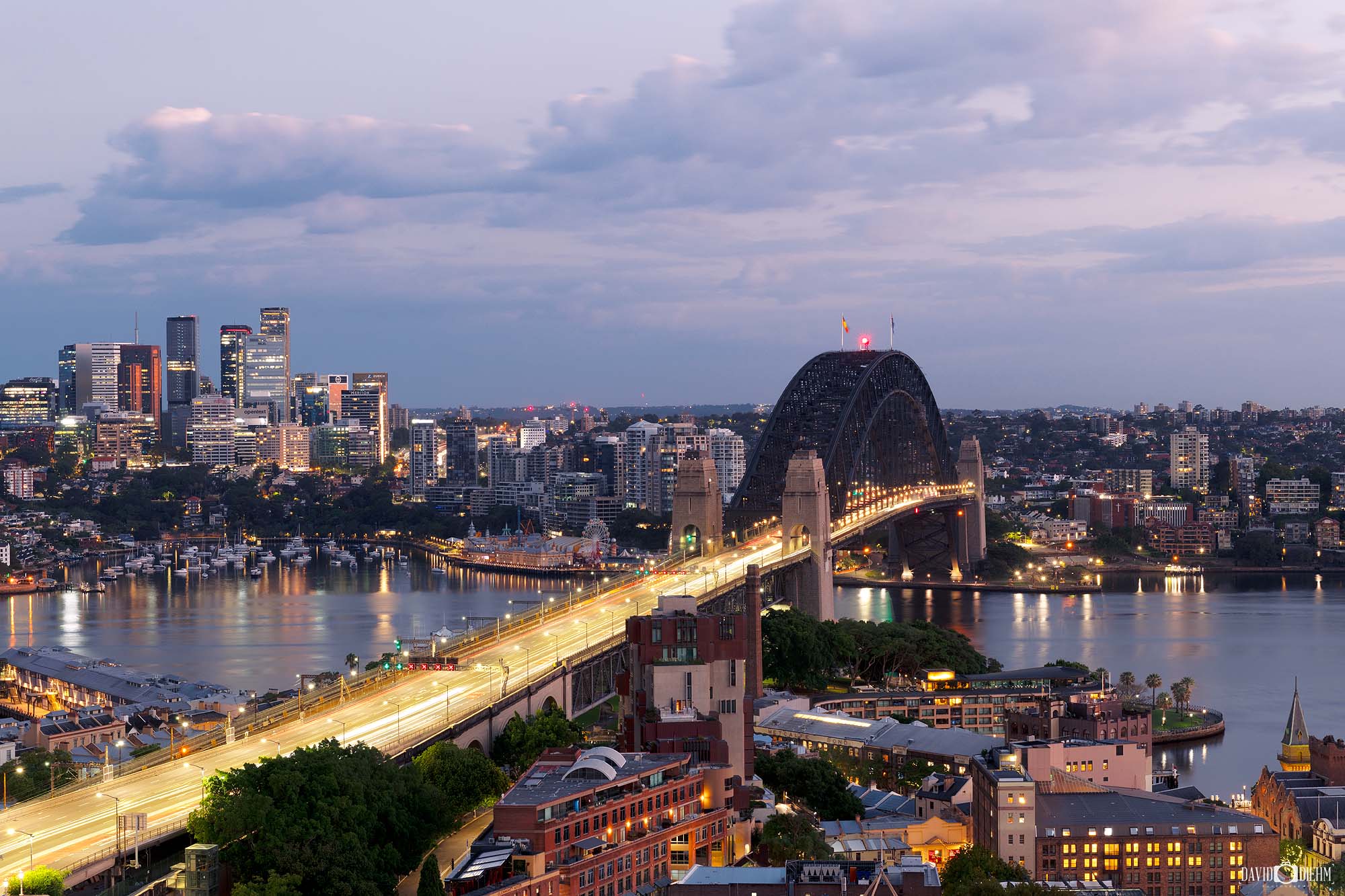 Long-exposure photograph of cars creating light trails across the Sydney Harbour Bridge at night with city lights in the background.
