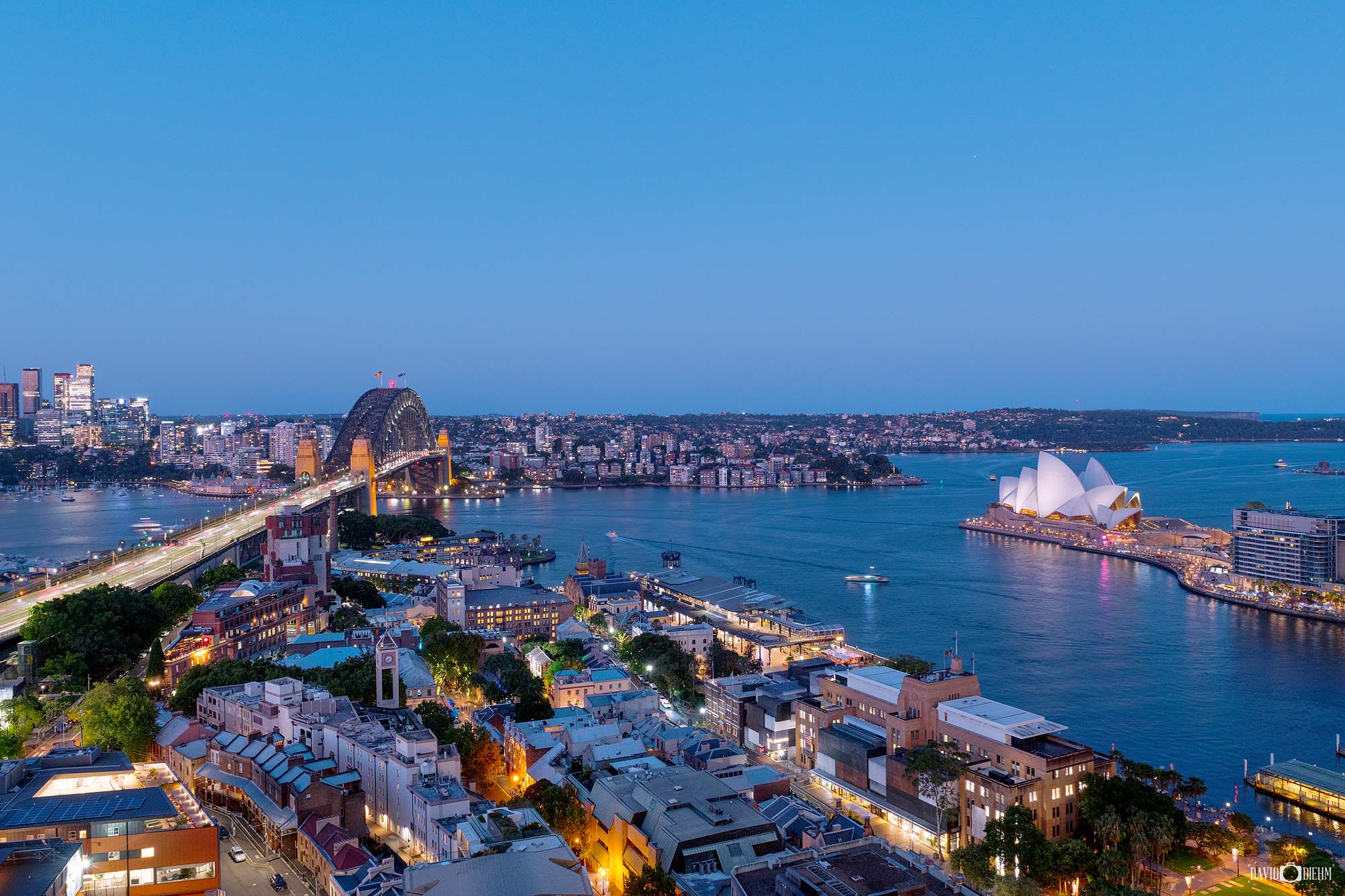 Photograph of Sydney Harbour at twilight with glowing city lights, waterfront reflections, and skyline views during the evening.