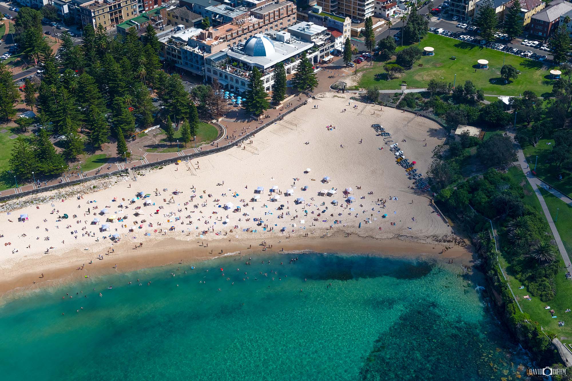Aerial photograph of Coogee Beach in Sydney, Australia, showing the curved shoreline, ocean pools, and coastal reserve from above.