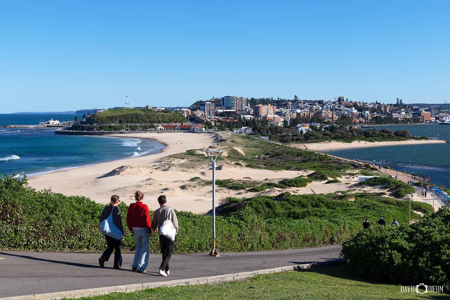 Visitors enjoying the views from Nobbys Headland Walk overlooking Nobbys Beach, Newcastle Harbour and the Newcastle skyline.
