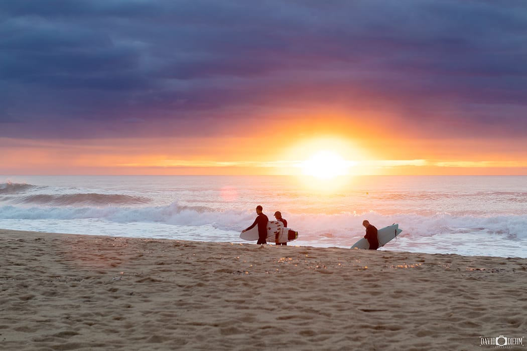 Surfers walking onto Dudley Beach at sunrise with waves breaking along the Lake Macquarie coastline