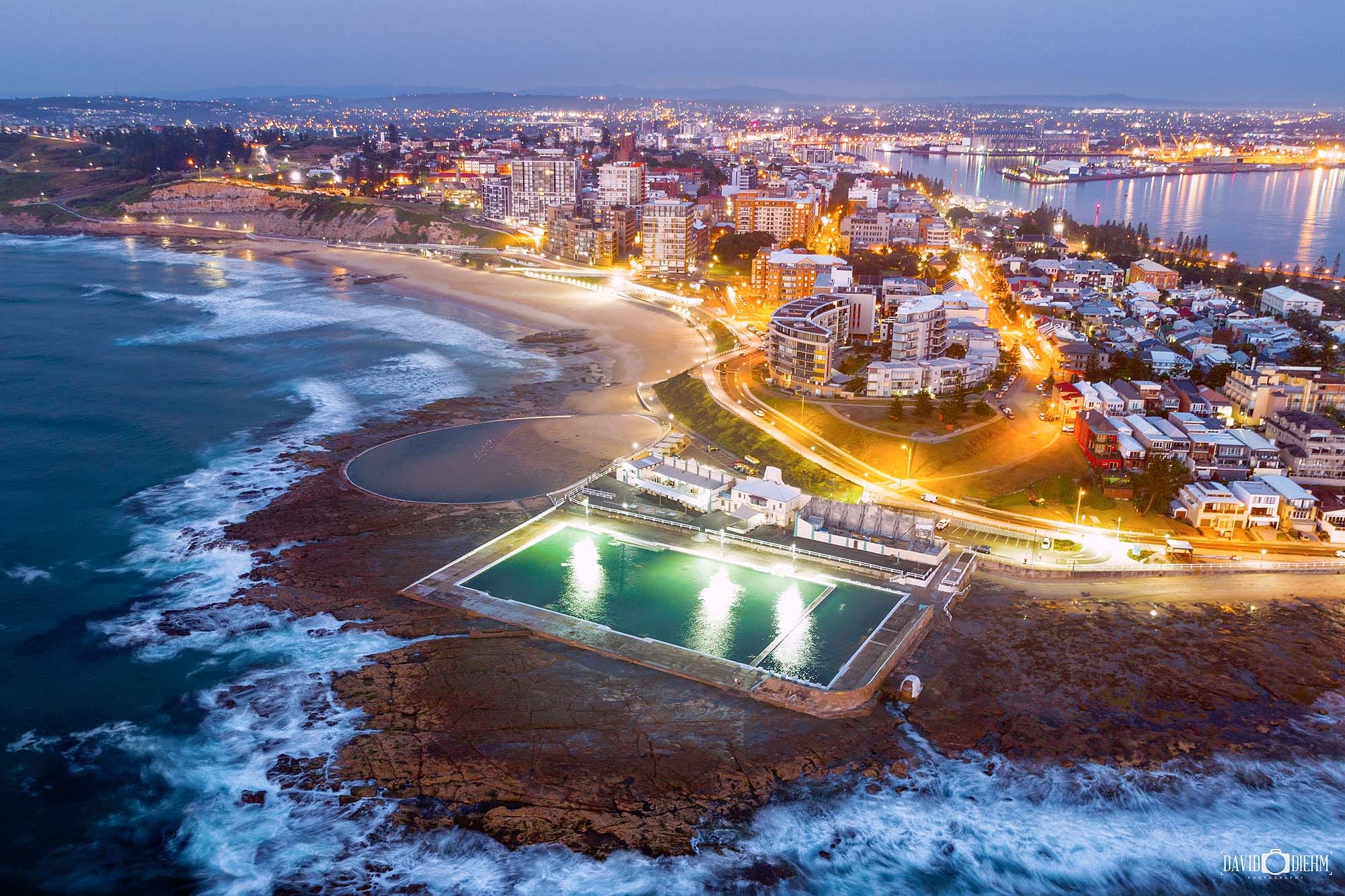 Aerial view of Newcastle Ocean Baths and Newcastle city skyline illuminated at dusk, Newcastle NSW Australia