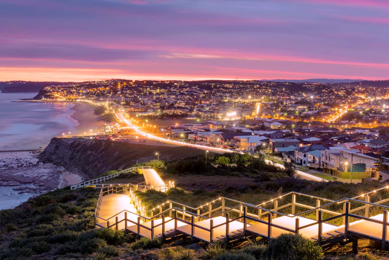 ANZAC Memorial Walk at twilight overlooking Bar Beach and Merewether, with illuminated boardwalk, city lights and colourful pink and purple sunset skies across Newcastle.