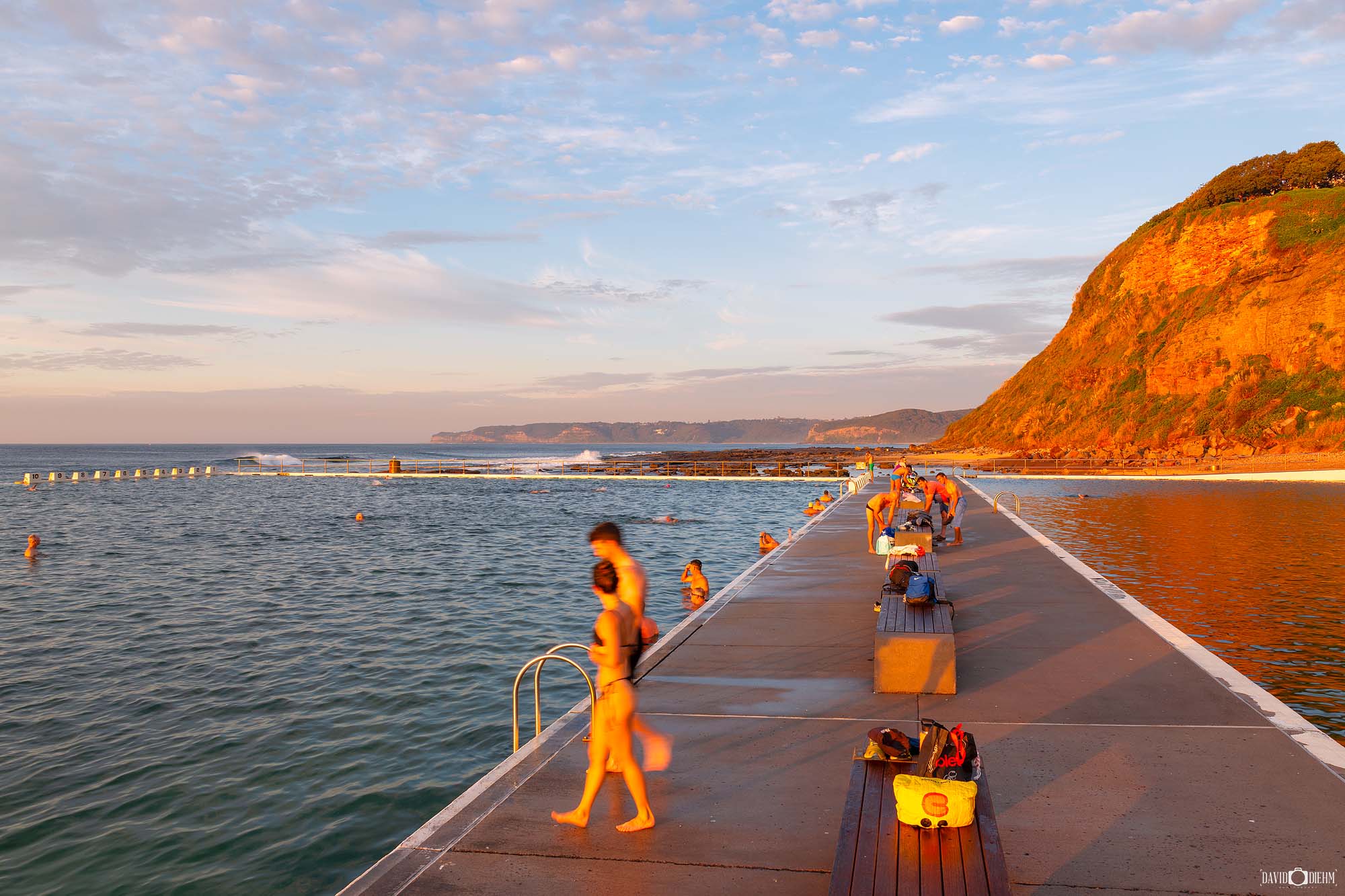 People enjoying a sunny morning at Merewether Ocean Baths with swimmers, walkers and visitors embracing Newcastle's coastal lifestyle.