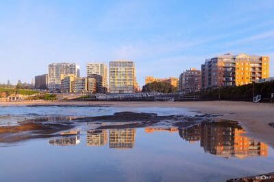 Sunrise reflected in the wet sand at Newcastle Beach with city buildings illuminated by golden morning light.
