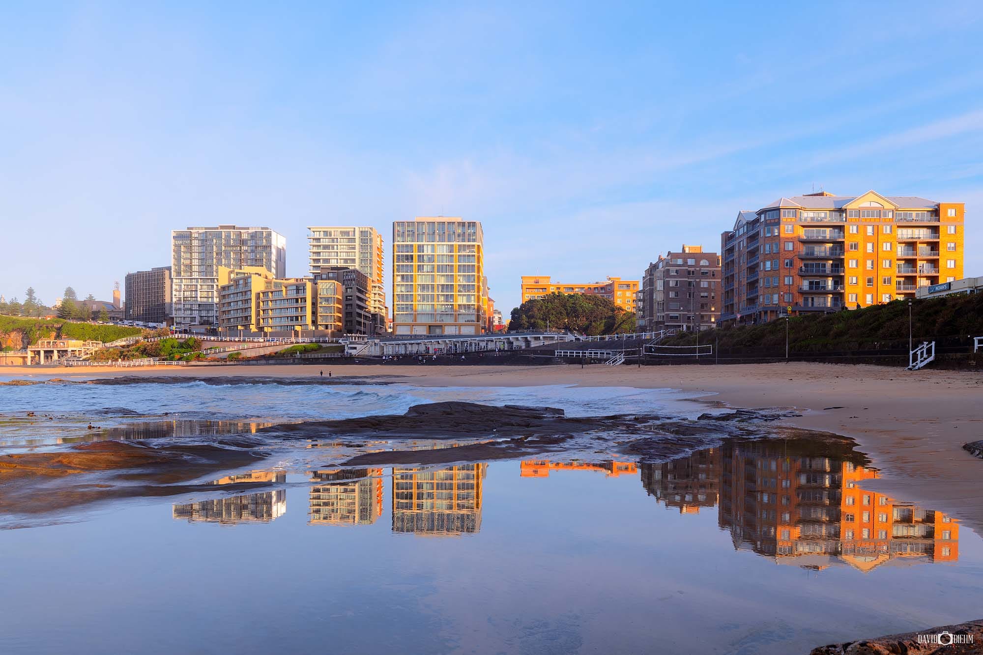Sunrise reflected in the wet sand at Newcastle Beach with city buildings illuminated by golden morning light.