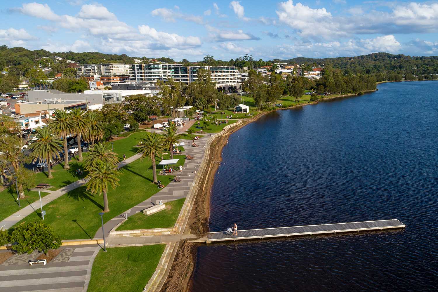 Aerial view of the Warners Bay foreshore on Lake Macquarie, showing the shared lakeside pathway, jetty, palm trees and waterfront parklands popular with walkers and cyclists.