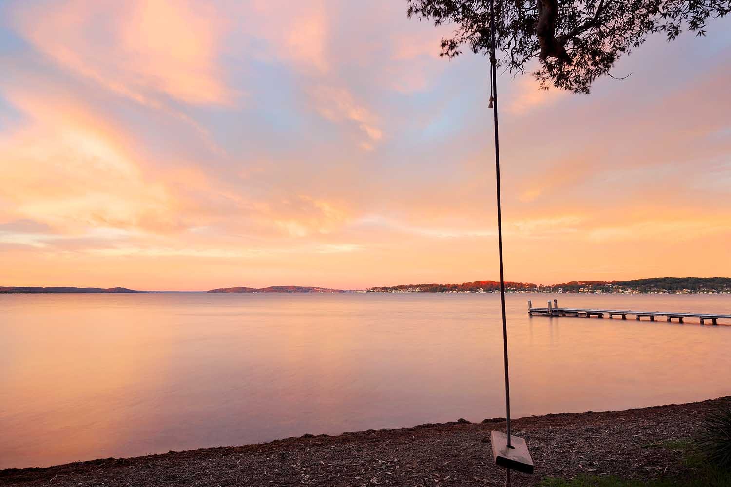 Sunset over Lake Macquarie at Green Point Reserve, featuring a lakeside swing and jetty silhouetted against soft pink and orange evening skies.