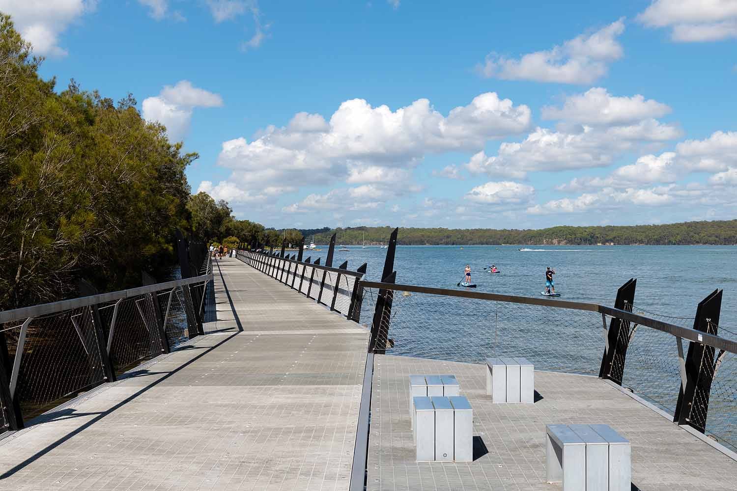 Visitors walking and paddleboarding beside the Red Bluff Boardwalk in Eleebana, Lake Macquarie, showcasing the elevated over-water pathway and scenic lakeside views.