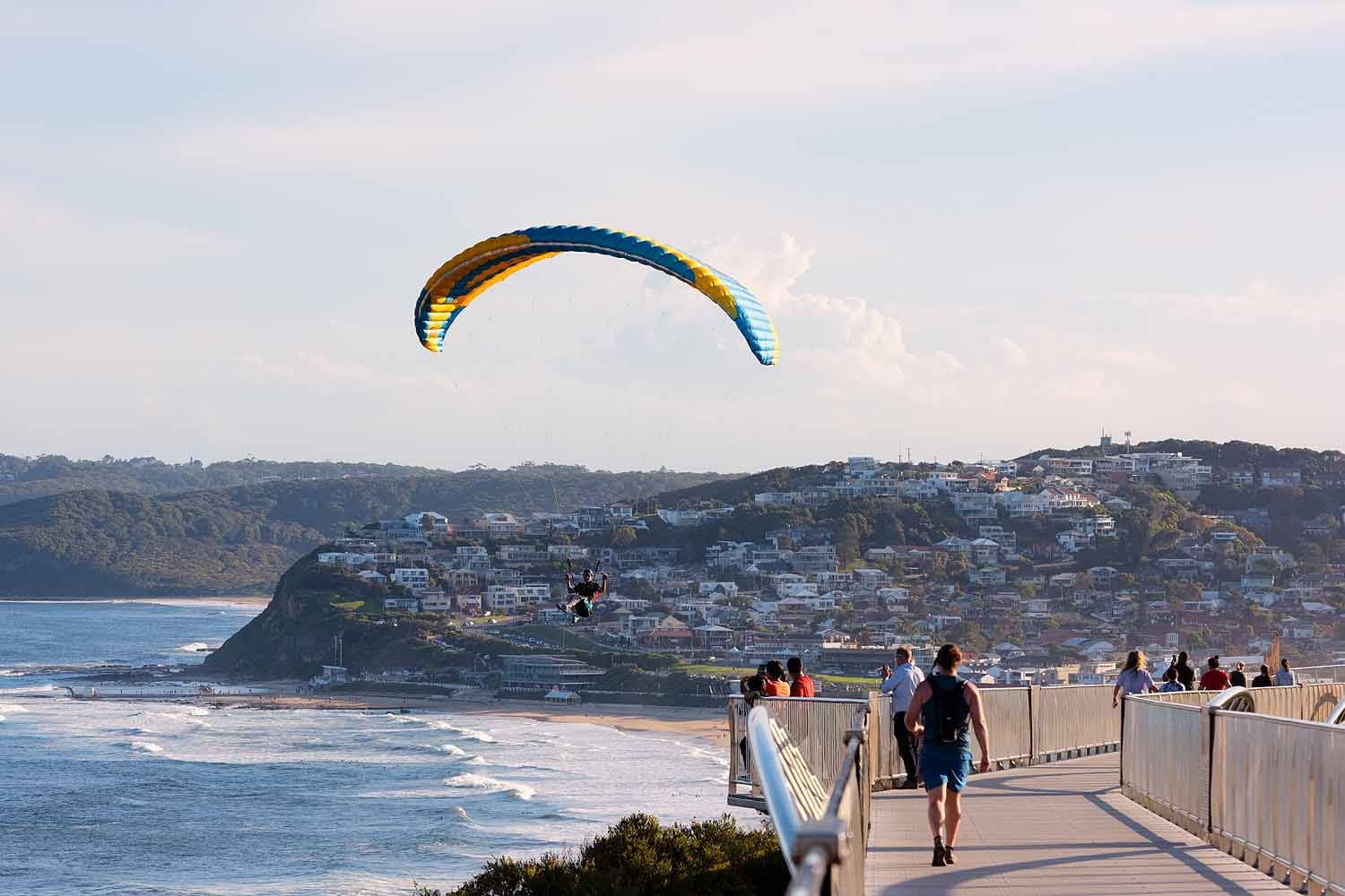 Paraglider soaring beside the Newcastle Memorial Walk above Bar Beach, with walkers enjoying panoramic coastal views towards Merewether and Glenrock.