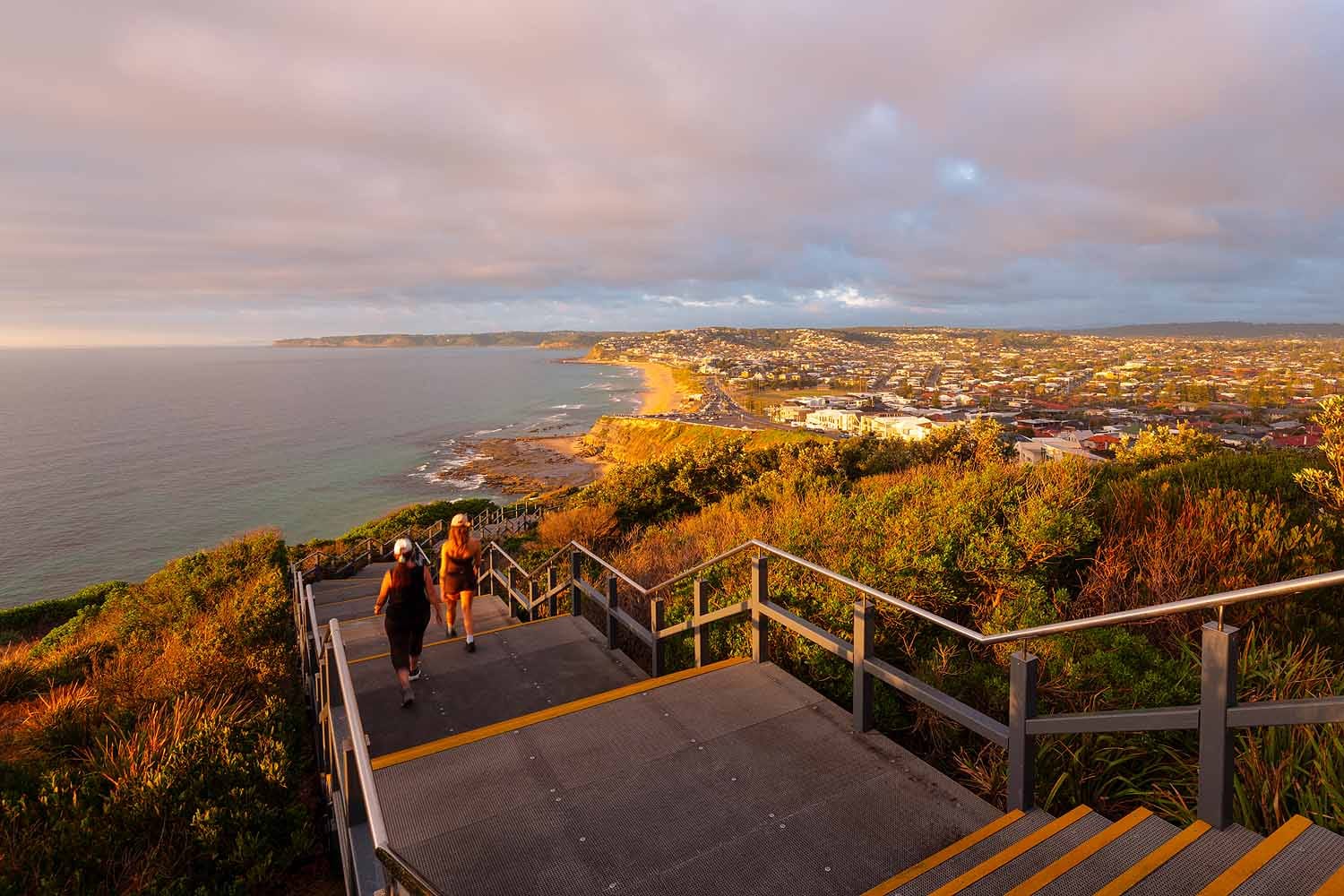 Sunrise view from the Newcastle Memorial Walk overlooking Bar Beach, Merewether Beach and the Newcastle coastline, with walkers descending the stairs in golden morning light.