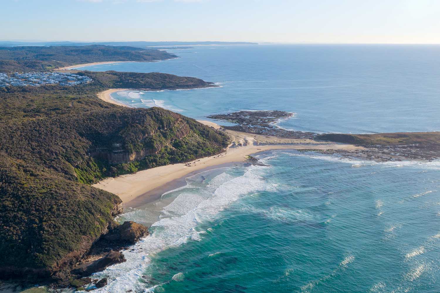 Aerial view of Ghosties Beach and the Munmorah coastline near Lake Macquarie, showcasing remote beaches, rugged cliffs, sea caves and turquoise ocean waters.