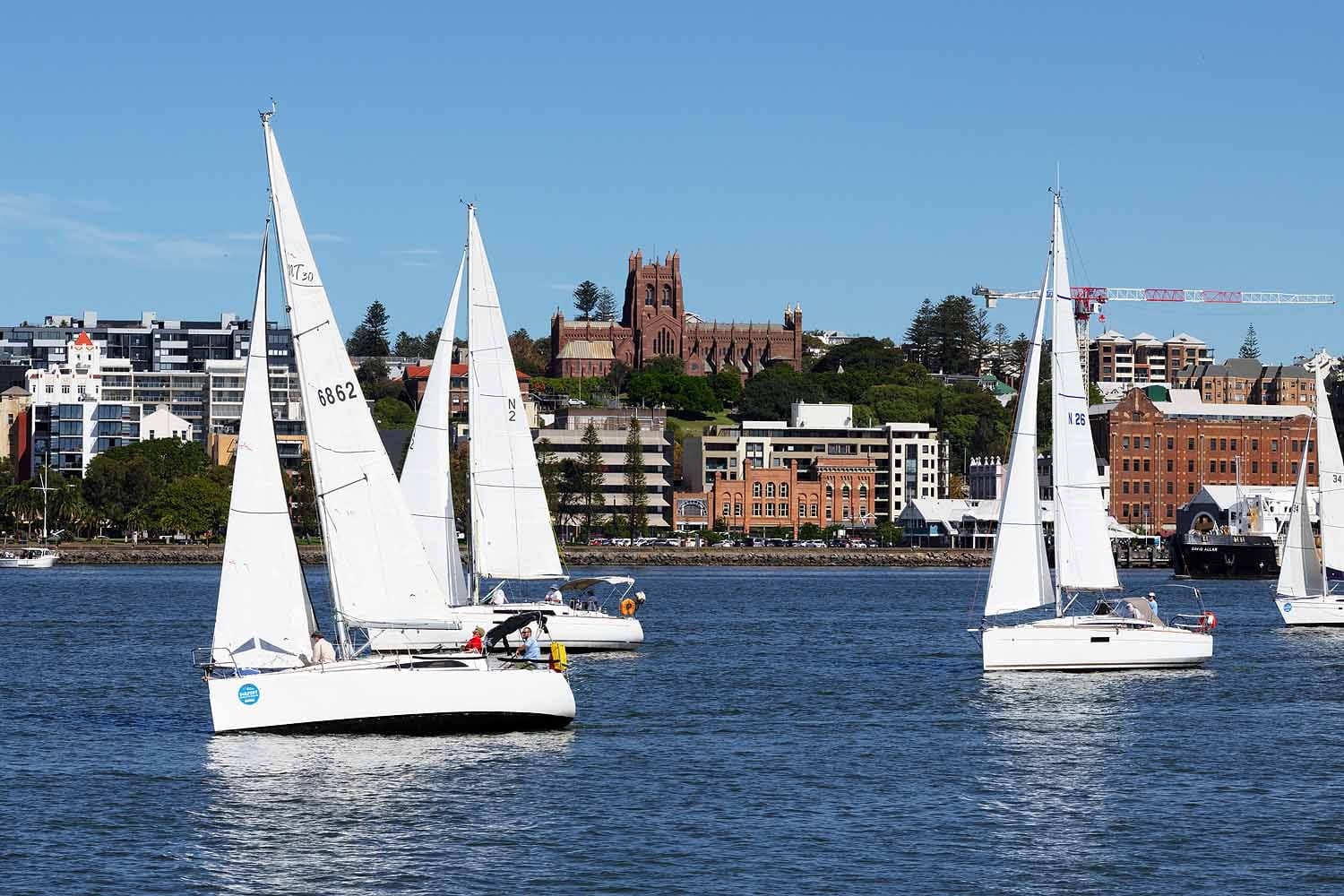 Yachts sailing across Newcastle Harbour with Christ Church Cathedral and the Newcastle city skyline in the background.