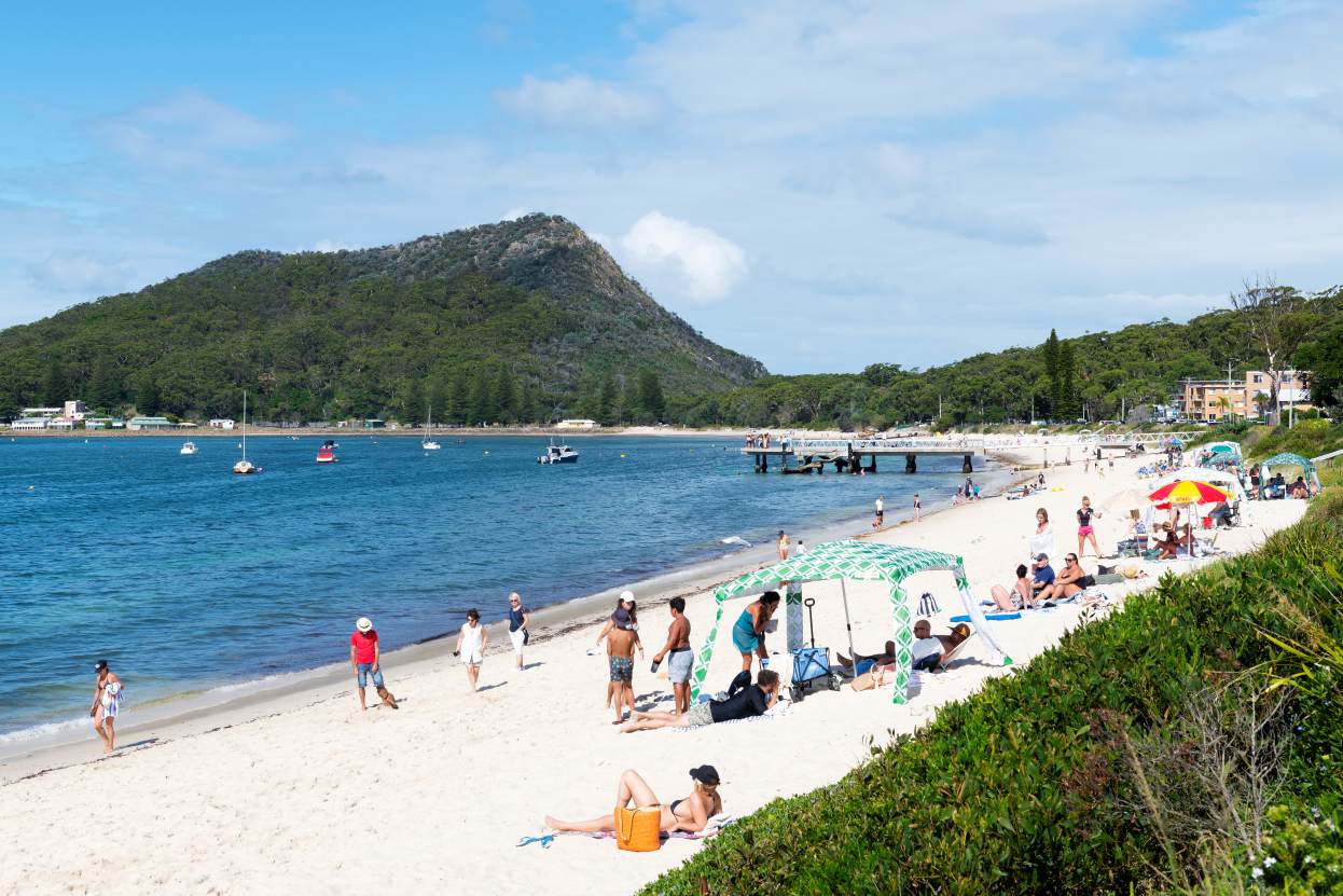 Visitors enjoying Shoal Bay Beach with Tomaree Head in Port Stephens NSW