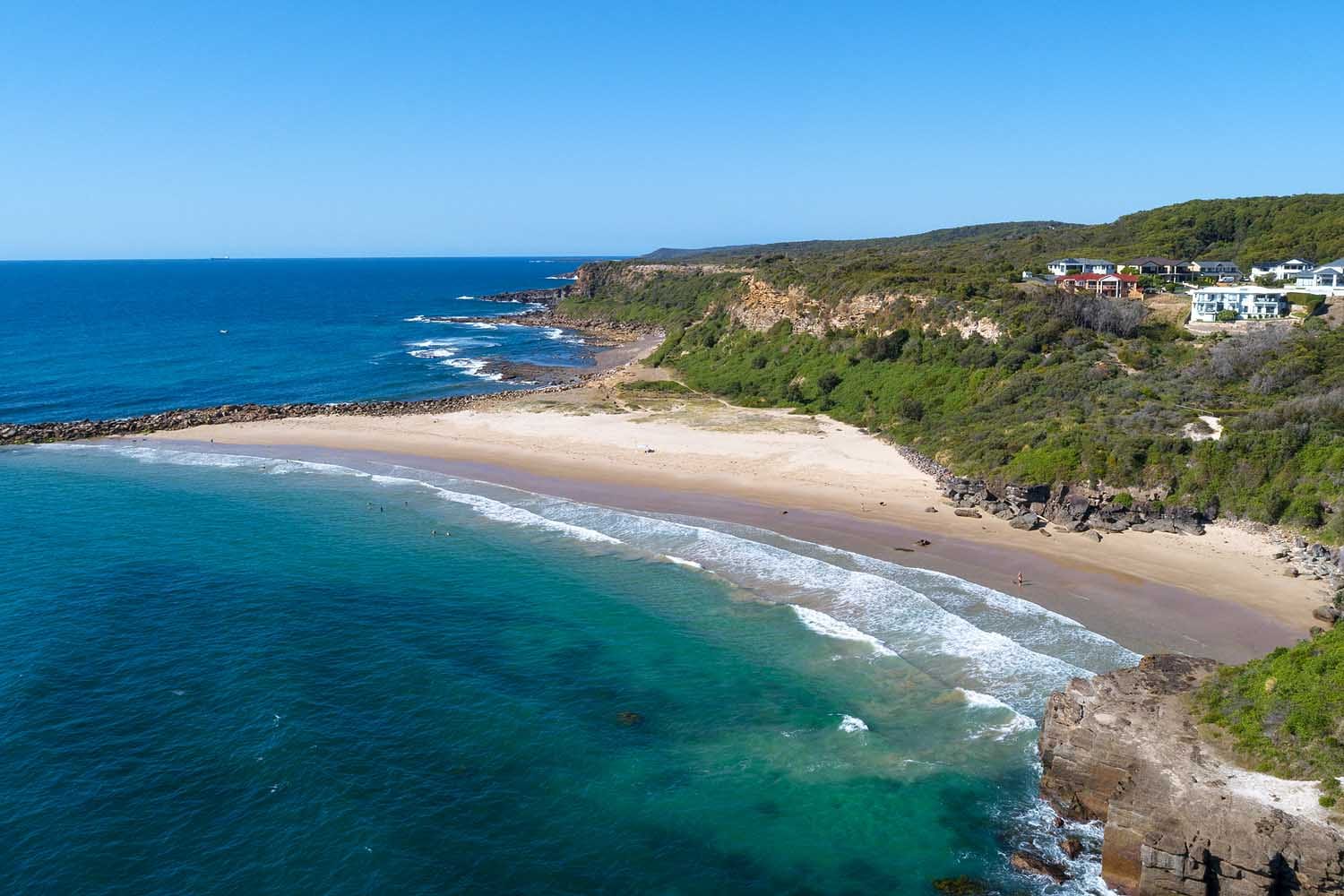 Aerial view of Spoon Rocks Beach near Lake Macquarie, featuring turquoise water, golden sand, rugged coastal cliffs and the sheltered bay within Munmorah State Conservation Area.