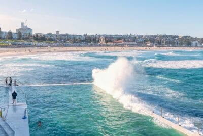 A wave crashes over Bondi Icebergs Ocean Pool with Bondi Beach, Bondi Pavilion and Sydney's iconic coastline in the background.