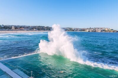 A powerful wave crashes over Bondi Icebergs Ocean Pool with Bondi Beach stretching across the background, Sydney NSW.