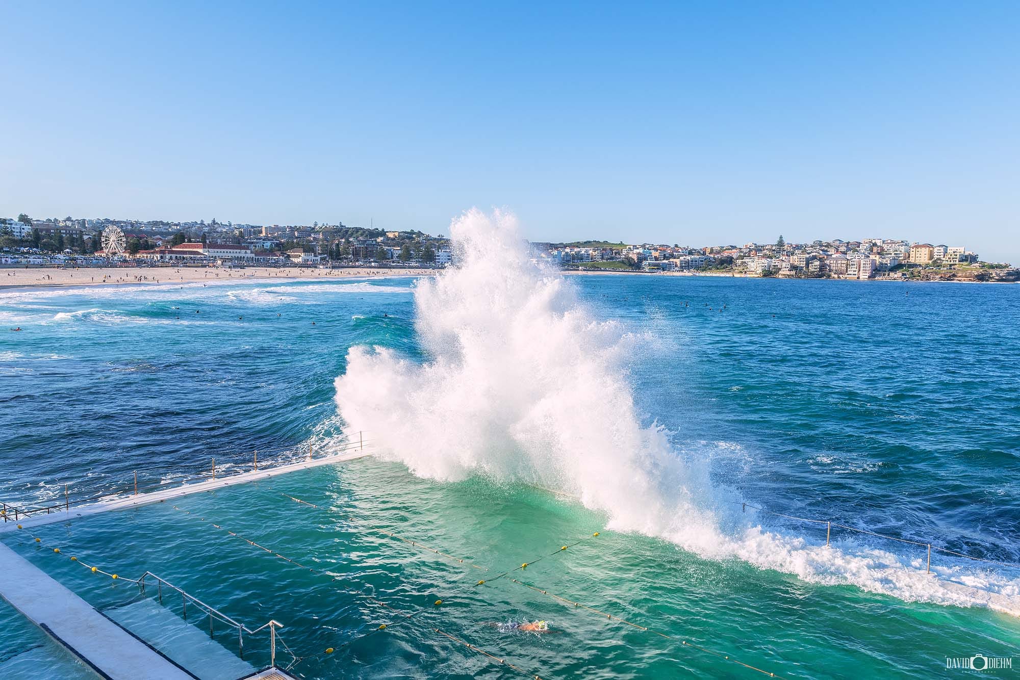 A powerful wave crashes over Bondi Icebergs Ocean Pool with Bondi Beach stretching across the background, Sydney NSW.