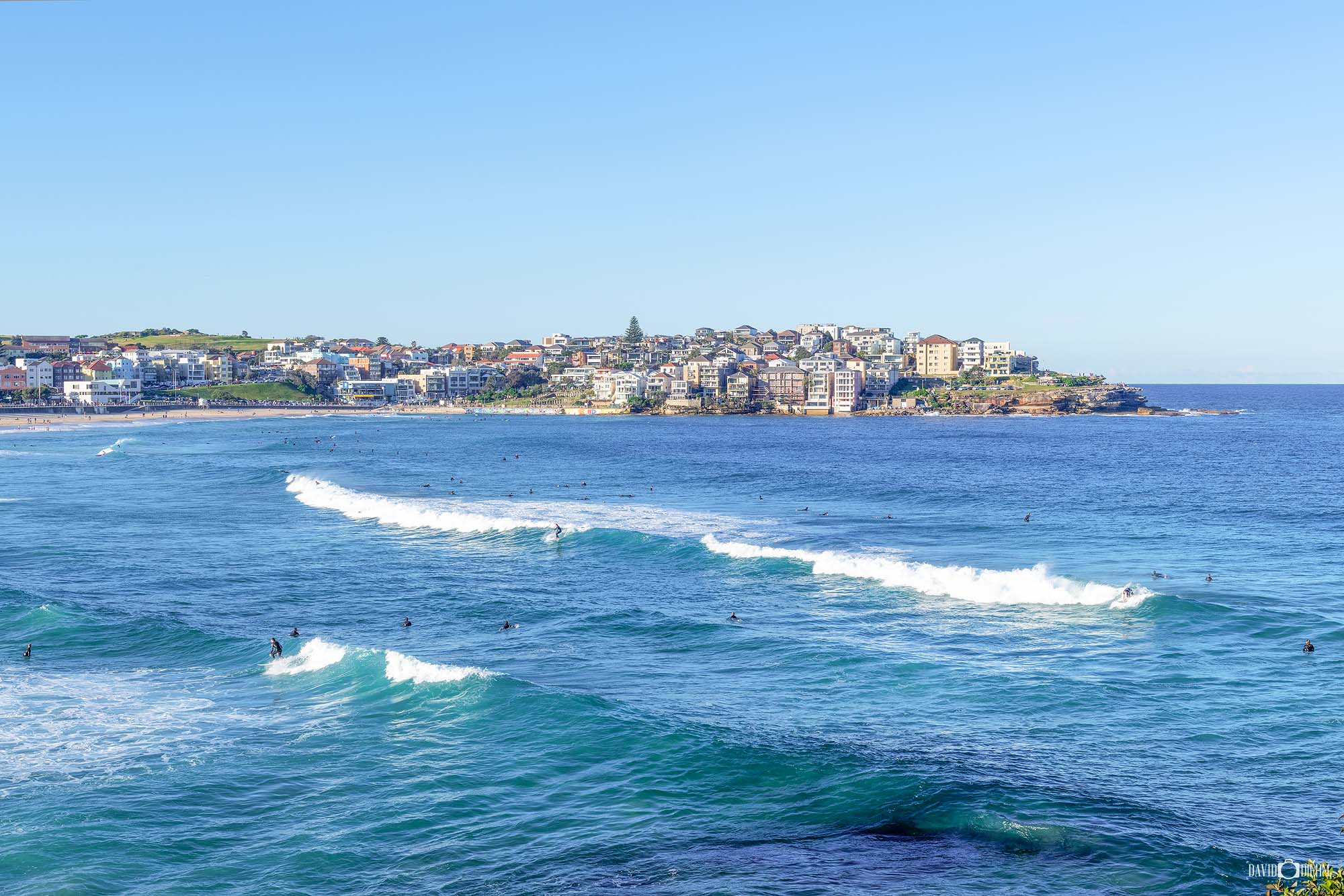 Clean surf lines roll towards Bondi Beach with surfers waiting for waves and the Bondi coastline in Sydney, NSW.