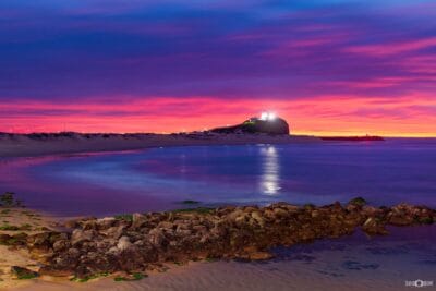 Vibrant sunrise over Nobbys Lighthouse reflected across calm water at Nobbys Beach in Newcastle NSW.