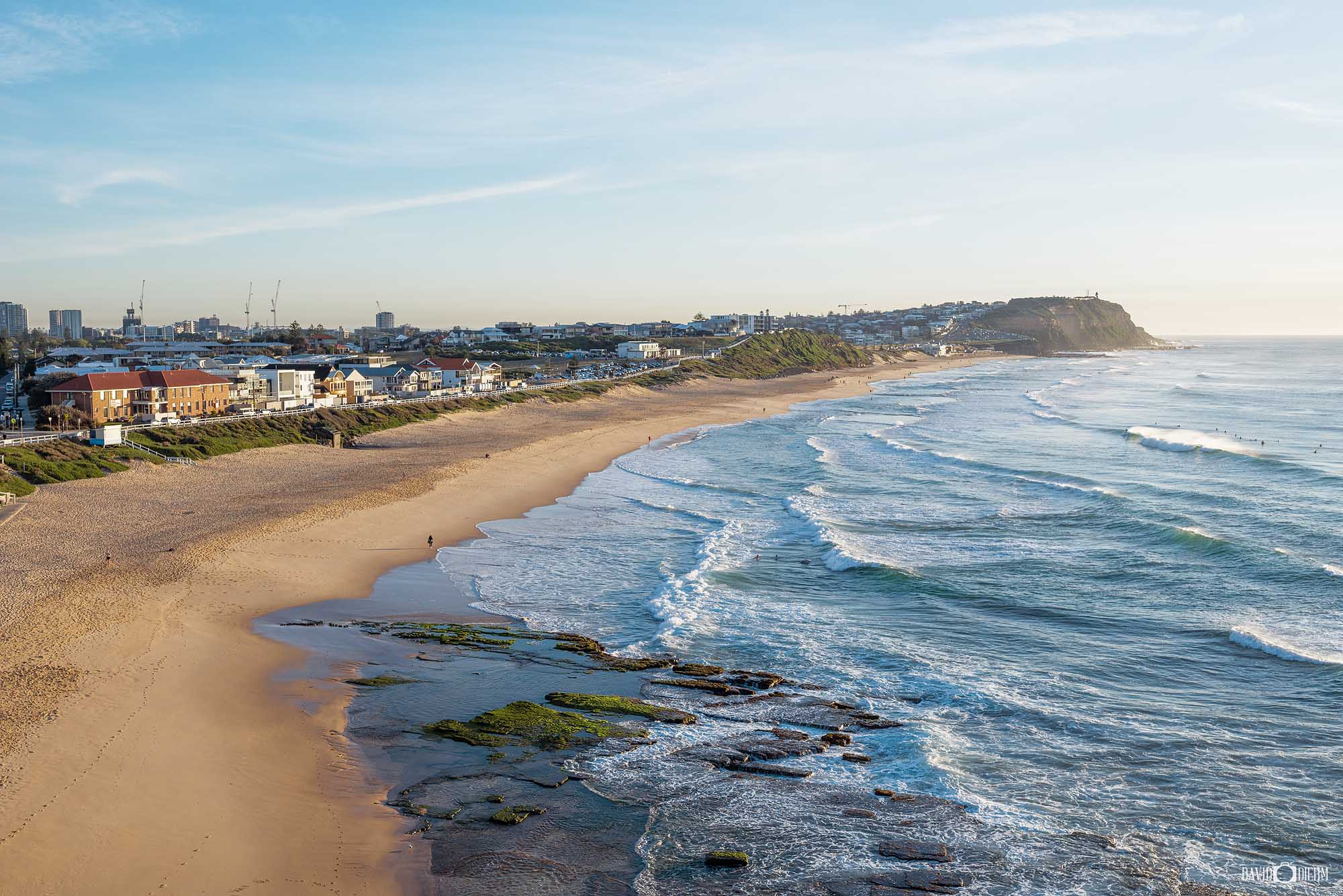 Aerial view along Merewether Beach in Newcastle NSW, with waves rolling onto the sandy coastline and the headland in the distance.