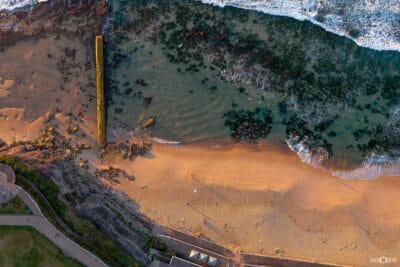 Aerial view looking directly down over Bar Beach in Newcastle NSW, with warm light across the sand, rock pool and ocean.