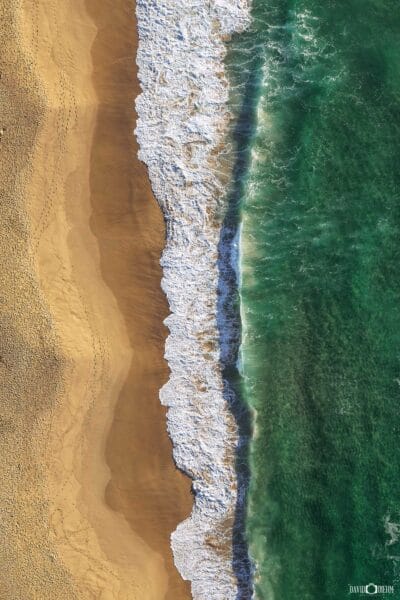 Aerial photograph of emerald-green ocean waves breaking onto golden sand, creating beautiful coastal patterns along the shoreline.