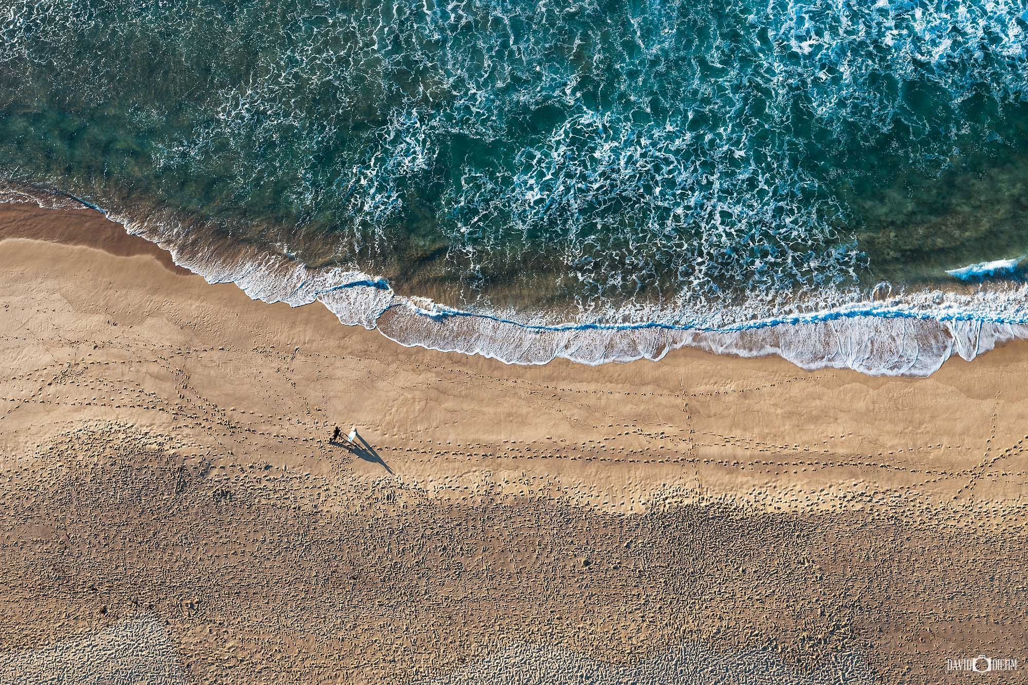 Aerial view looking down over Merewether Beach in Newcastle NSW, with a surfer walking beside the shoreline and footprints across the sand.