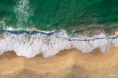 Aerial photograph of emerald-green ocean waves gently embracing the golden shoreline, with intricate white surf patterns stretching across the sand.