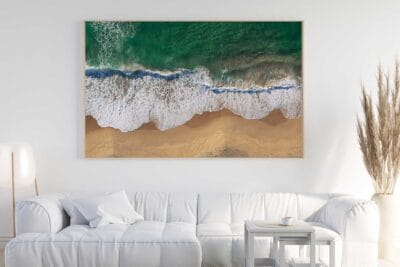 Shoreline Embrace aerial coastal photography print displayed in a natural timber frame above a white sofa in a bright, contemporary living room.