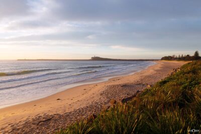 Stockton Beach bathed in warm morning sunlight, with gentle waves rolling onto golden sand and Nobbys Headland visible across the water in Newcastle NSW.