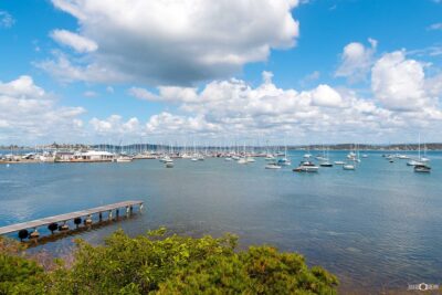 Sailboats anchored at Belmont on Lake Macquarie beneath fluffy white clouds and a brilliant blue sky.