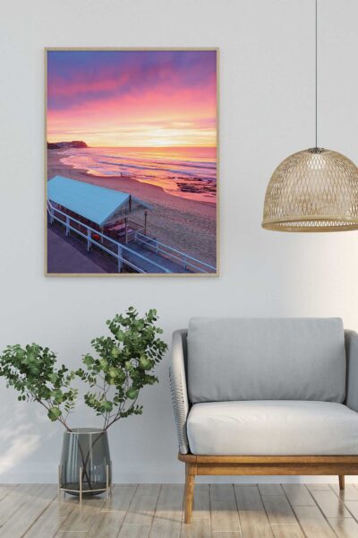 Portrait framed photograph of Merewether Beach Pavilion at sunrise, featuring vibrant pink and golden skies, displayed above an armchair in a coastal-inspired interior.