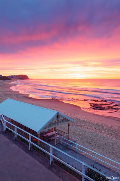 Portrait photograph of Merewether Beach Pavilion in Newcastle beneath a spectacular pink, purple and golden sunrise reflecting across the ocean.