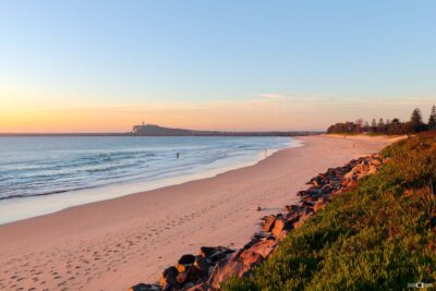 Stockton Beach glowing in warm golden light, with soft waves, pink-tinted sand and Nobbys Headland in the distance across Newcastle Harbour.