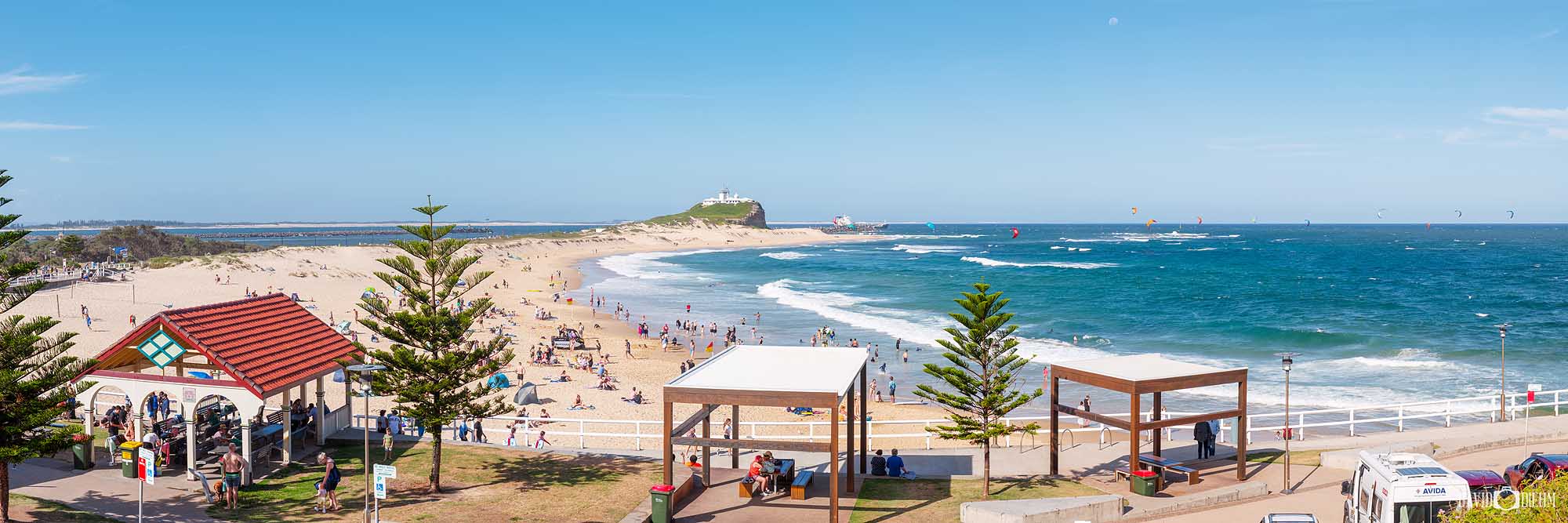 Panoramic view of Nobbys Beach in Newcastle NSW on a vibrant summer day, with swimmers, beachgoers, colourful kitesurfers and Nobbys Lighthouse in the distance.