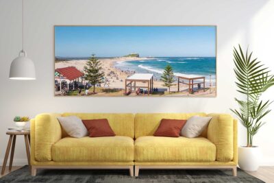 Nobbys Summer in Full Swing panoramic beach photography print displayed in a natural timber frame above a yellow sofa in a contemporary living room.